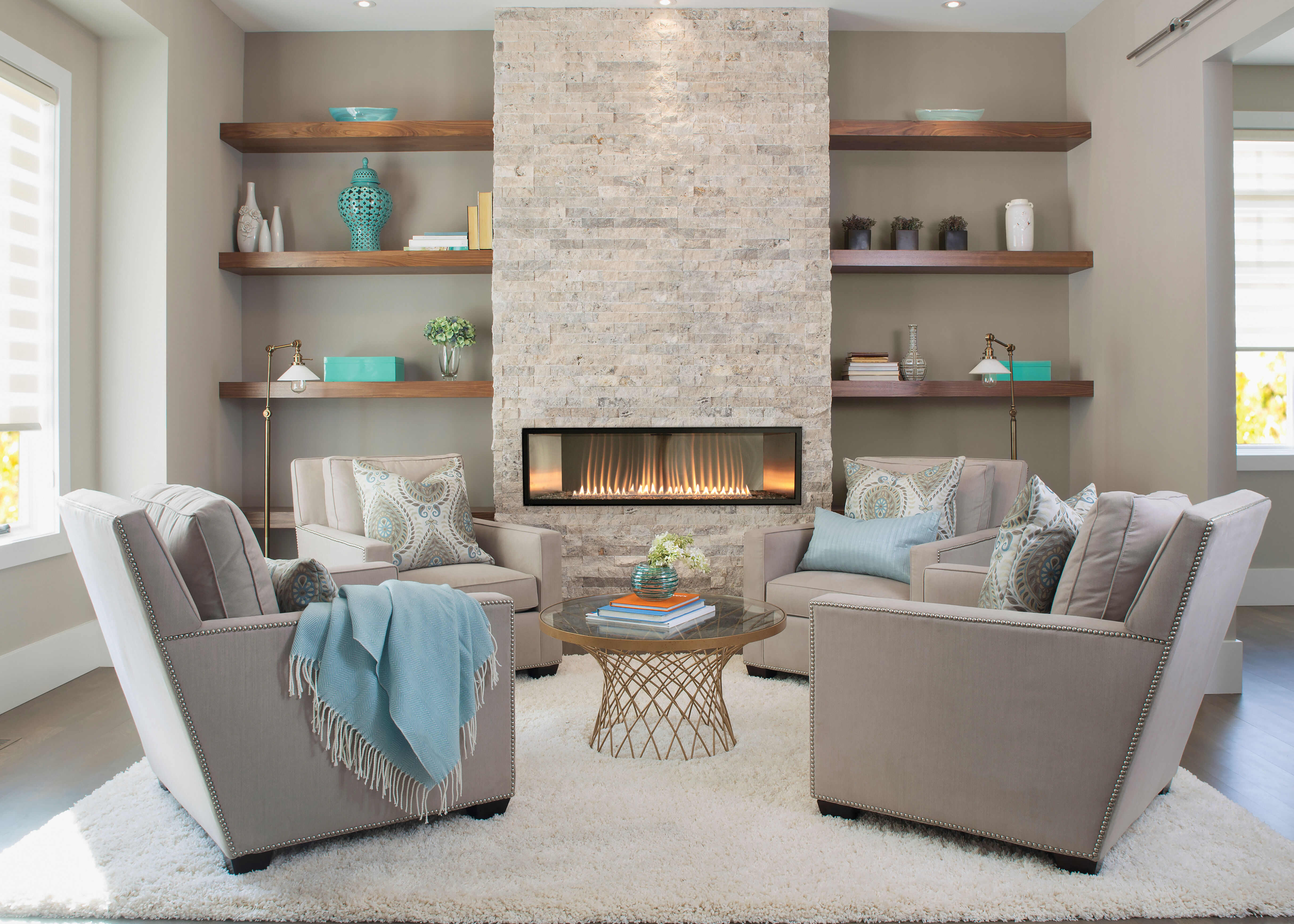 A contemporary living room with a large, natural stone hearth and linear gas fireplace, built-in shelving with blue accent pieces, and gray furniture with blue blankets and pillows.