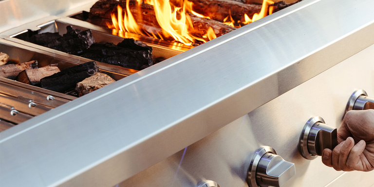 A close-up of a person's hand turning a silver control knob on a stainless steel hybrid grill. Inside the grill, separate fuel trays contain charcoal and wood logs, with one section showing active orange flames.