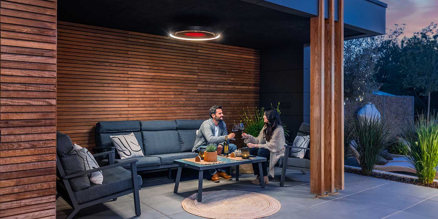A man and a woman toasting with two glasses of wine on an outdoor patio underneath a Bromic Eclipse Pendant patio heater that's mounted on the ceiling.