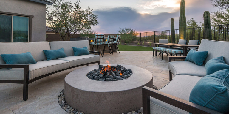 An outdoor patio space with a large, round concrete fire pit and two sectional outdoor couches