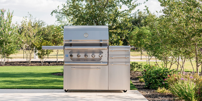 A large, professional-grade stainless steel gas grill with two side cabinets and locking wheels, sitting on a tiled patio. The grill is in an expansive backyard that has artificial turf and landscaping. Behind it is a black metal fence and green trees.