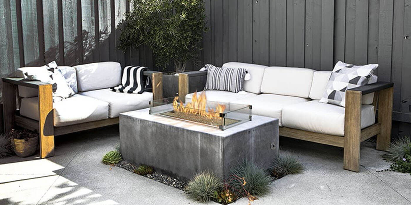 An outdoor living space with two couches and a concrete, rectangular gas fire pit with a glass wind guard  An outdoor living space with two couches and a concrete, rectangular gas fire pit with a glass wind guard