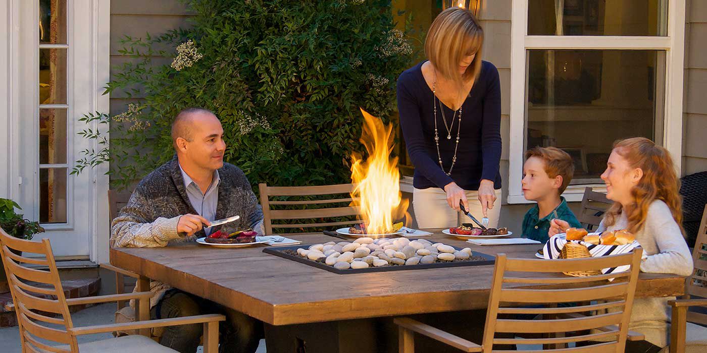 A family sitting around an outdoor gas fire pit dining table 