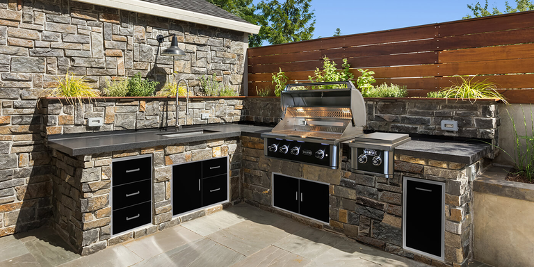 An L-shaped outdoor kitchen with a rugged stone base and dark granite countertops features an open grill, matching ice chest, and black-accented storage. Behind the continuous countertop, a thriving herb garden planter runs under a rustic gooseneck barn light.