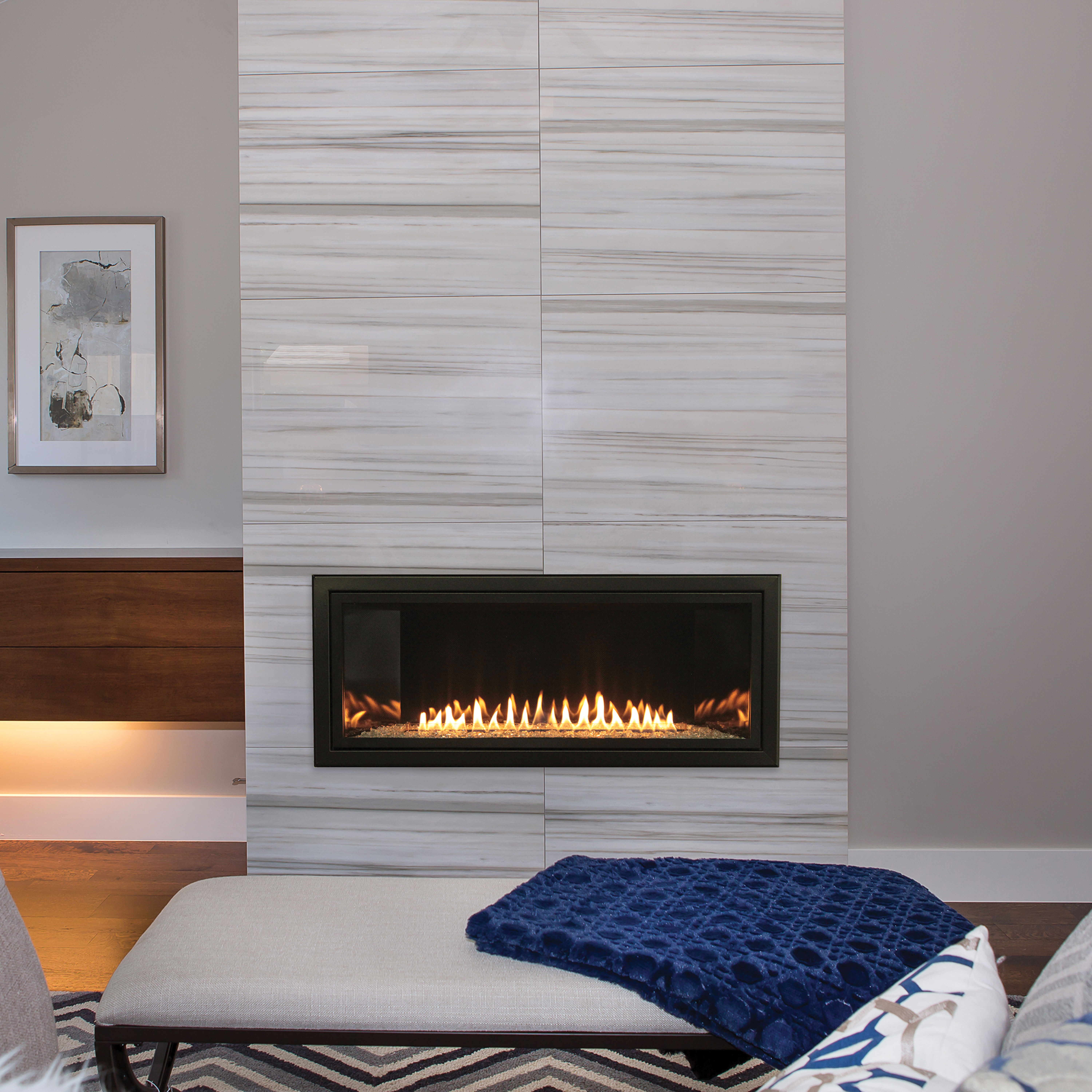 The Empire Contemporary Boulevard Ventless Gas Fireplace installed on a white, textured wall with a grey, geometric rug, a white ottoman and a blue accent blanket.