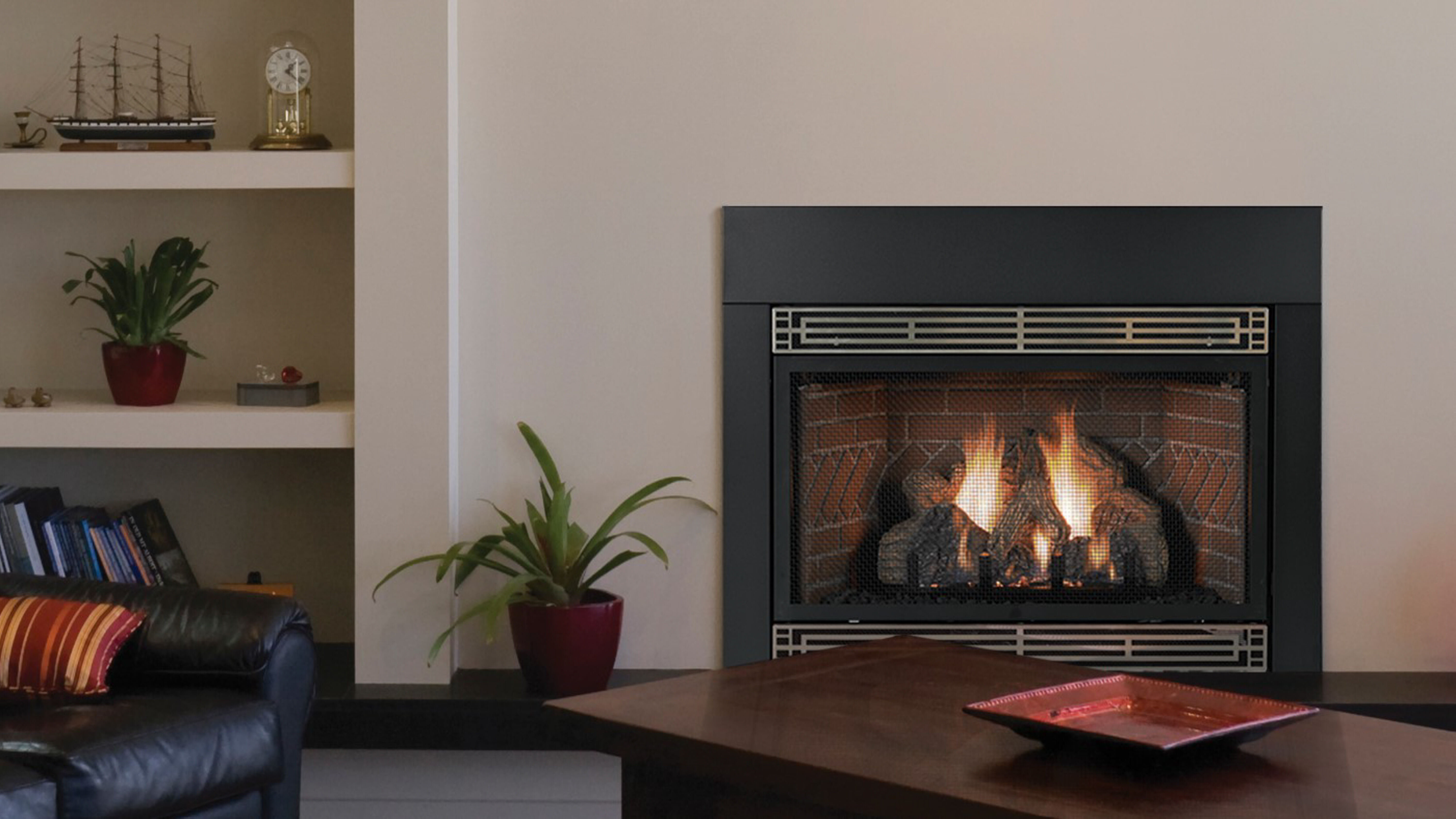 A traditional living room with a black leather couch, a dark wood coffee table, potted plants, built-in shelves, and a large, square fireplace with black trim and a black mesh safety screen.