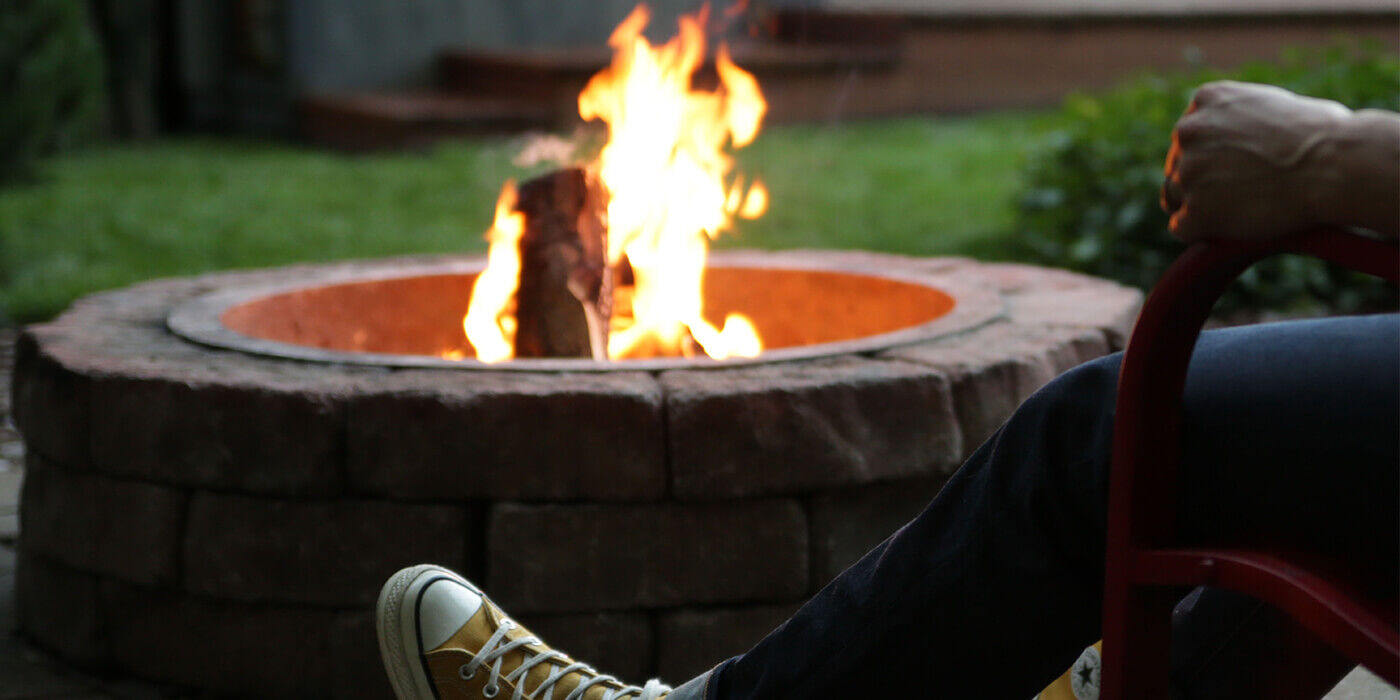 A large natural stone wood burning fire pit with a person sitting in a chair beside it