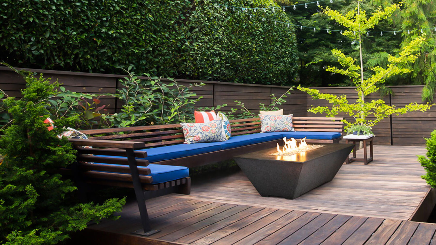 An outdoor deck surrounded by lush green foliage with a large, L-shaped blue couch and a black gas fire table.