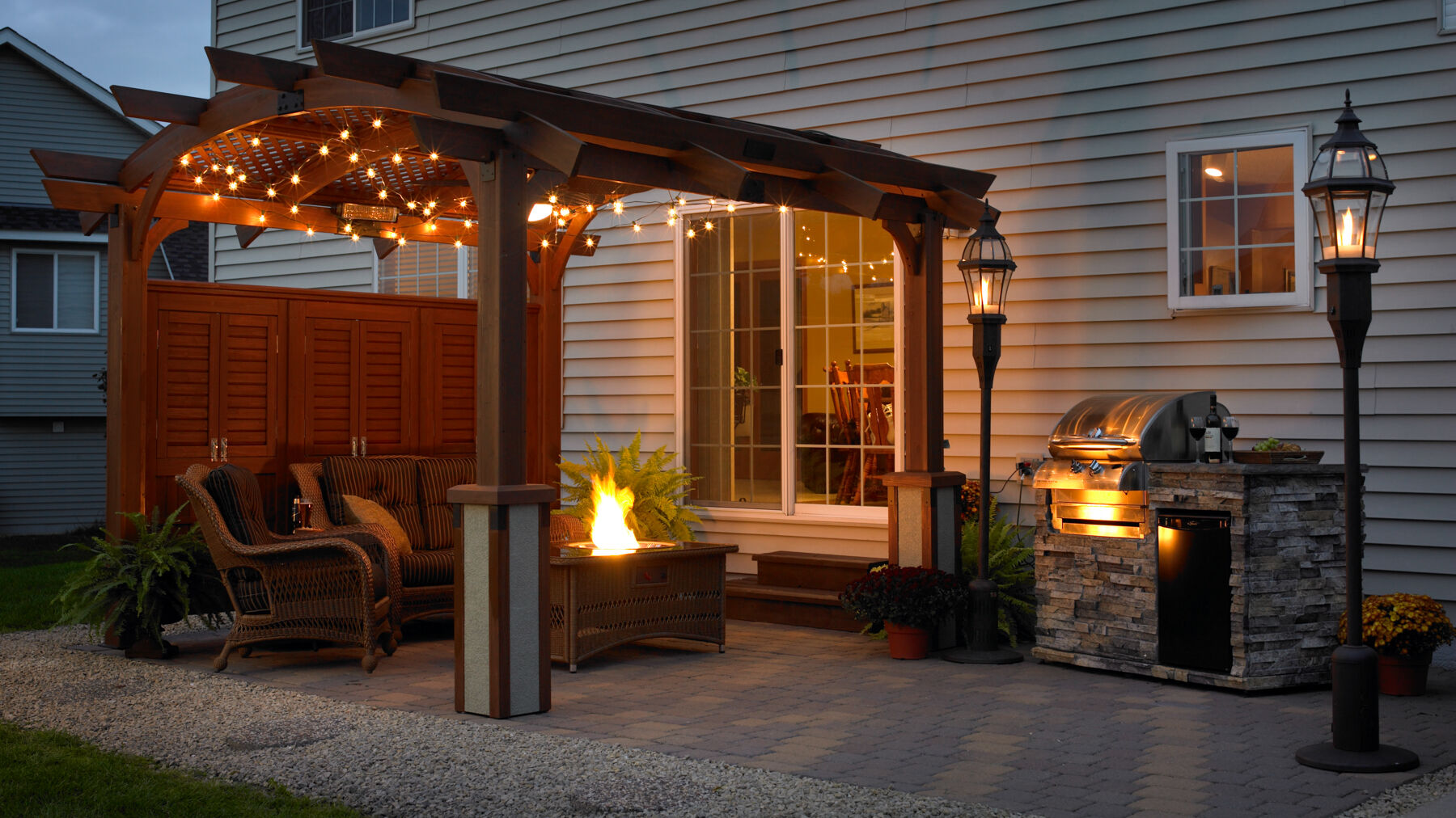 An outdoor patio space under a gazebo at night with fairy lights in the rafters, wicker lounge furniture, a gas fire pit table, a small kitchen island with a gas grill, and gas tiki torches.