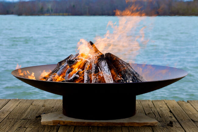The Asia Wood Burning Fire Pit by Fire Pit Art on top of a concrete slab, placed on a wooden deck with a lake and trees in the background.