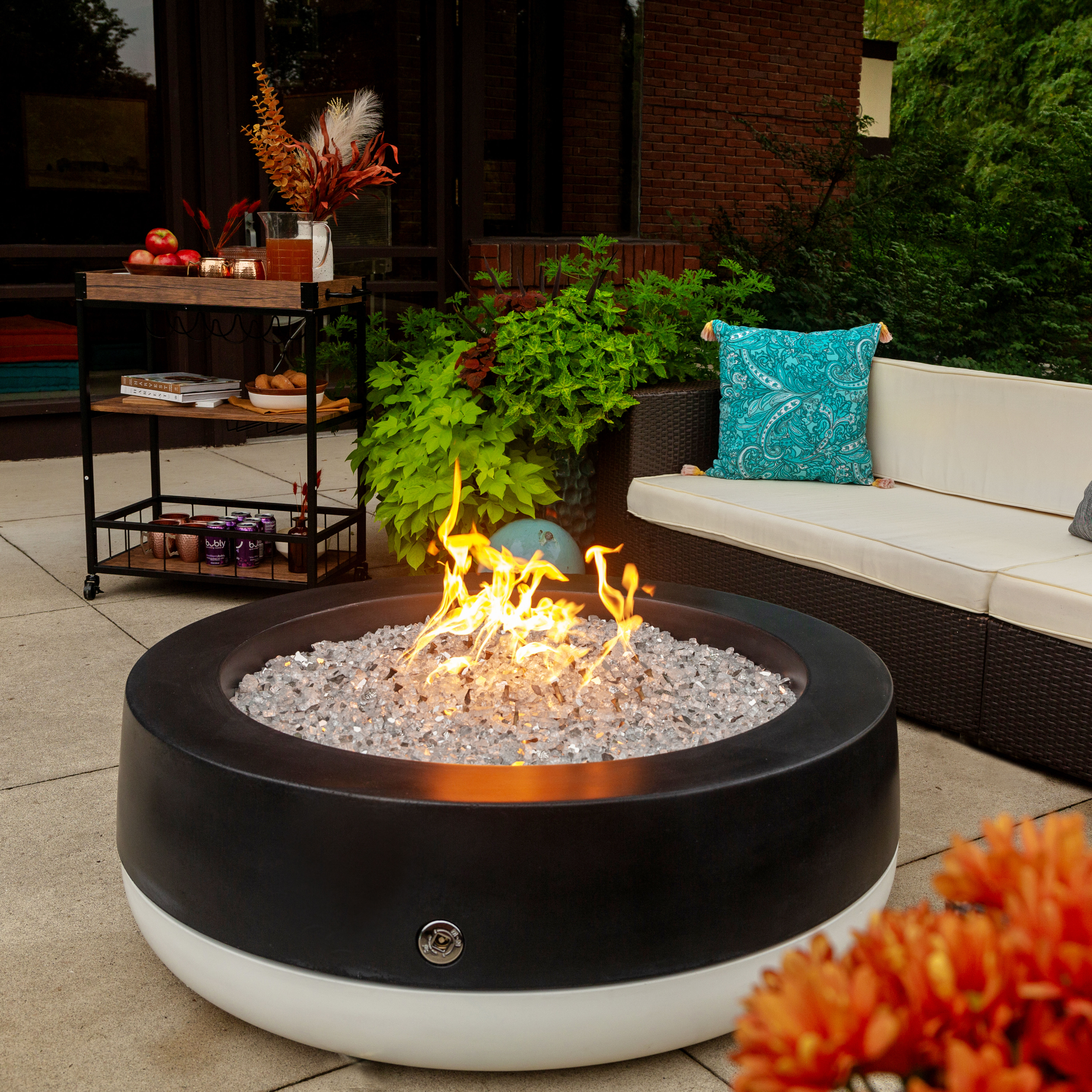 The FlameCraft Black and White Tondo Gas Fire Pit on a concrete patio with black and beige furniture, a wooden bar cart with refreshments, and lush green plants.