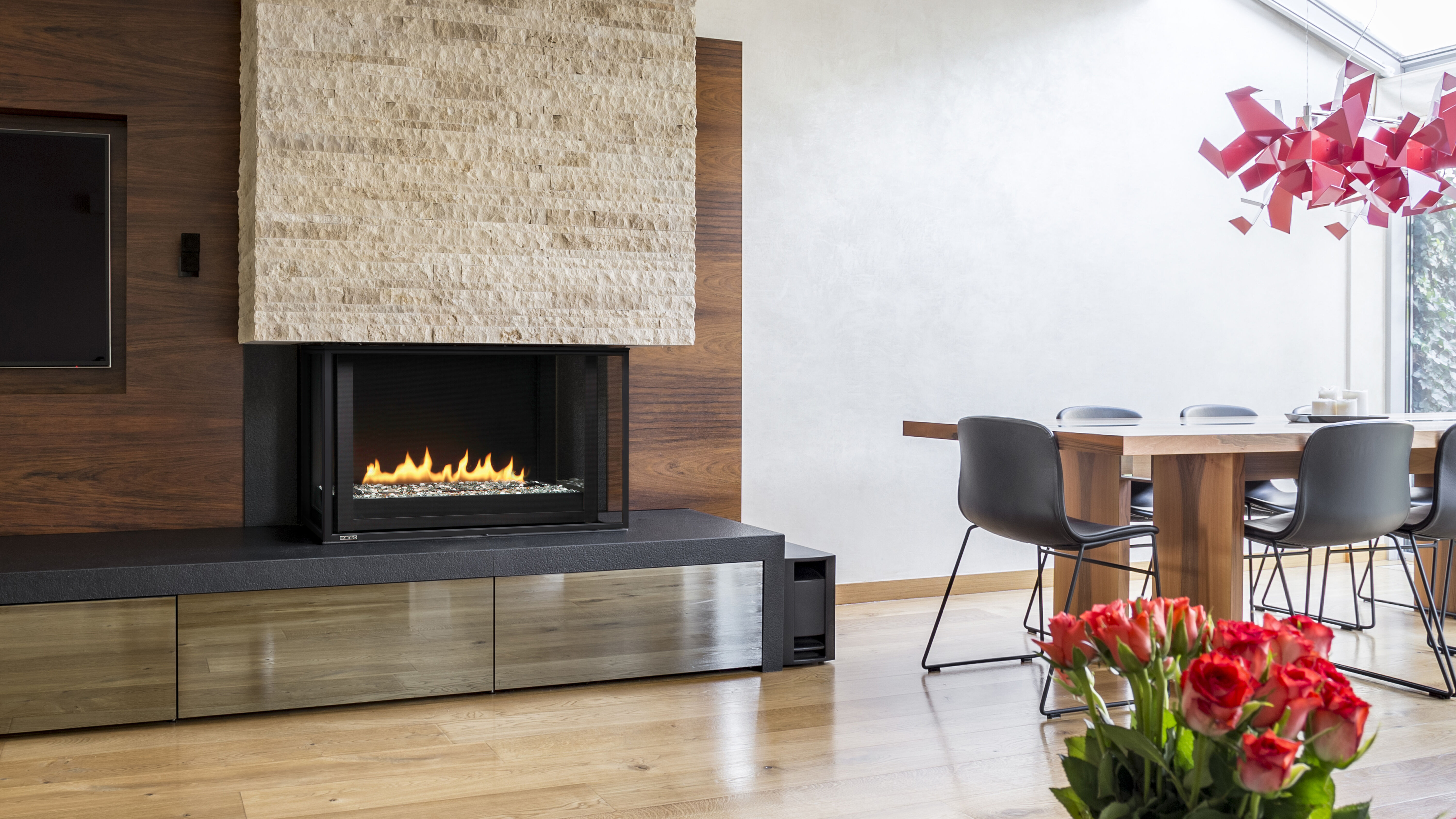 A modern, minimalistic living space with a large wooden dining table, eight black metal chairs, and a hearth finished with wooden planks, a natural stone mantel, and a linear gas fireplace.