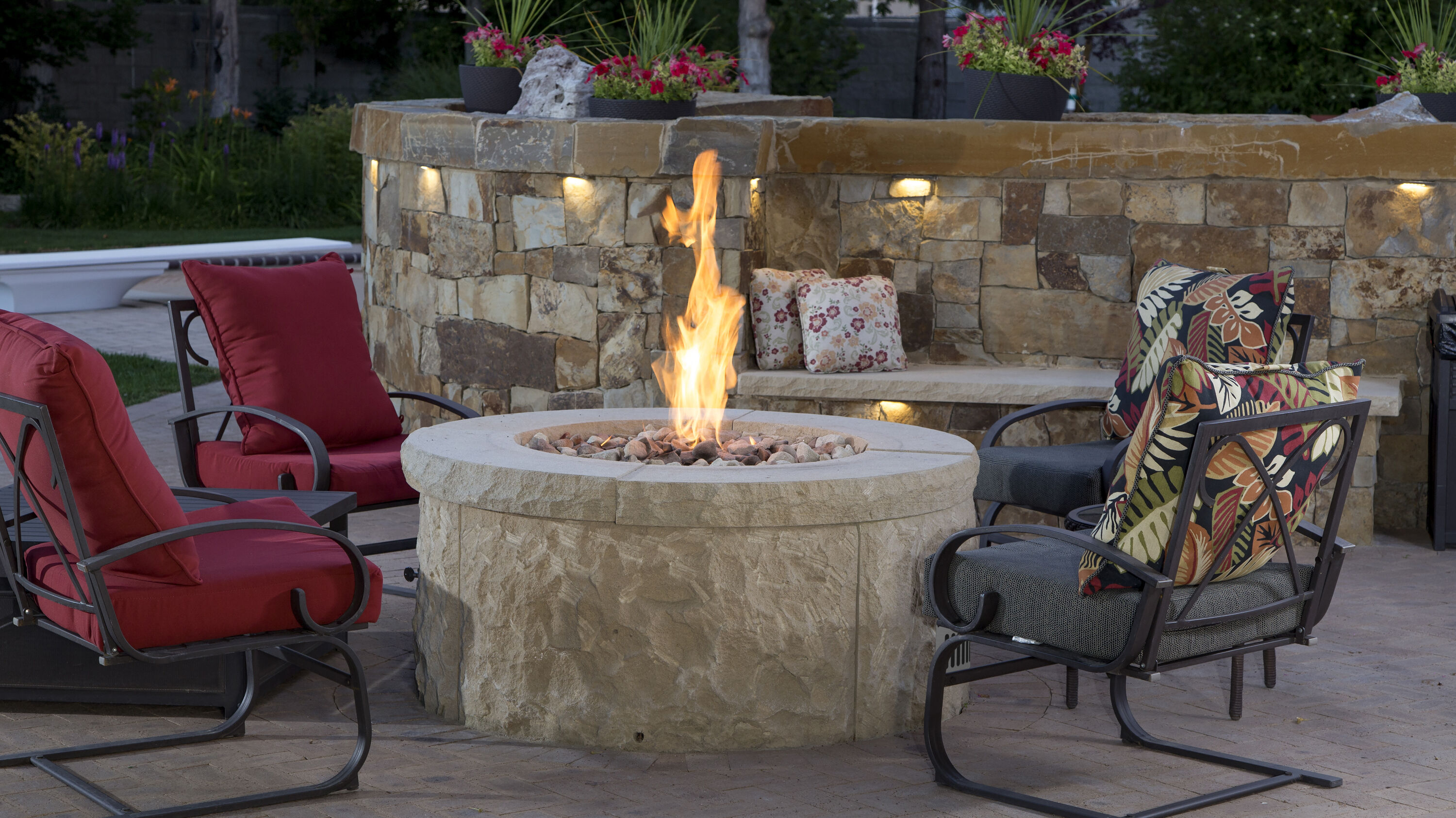 An outdoor patio with three red cushioned lounge chairs and a medium-sized, natural stone gas fire pit with multi-colored river rocks in the media bed.