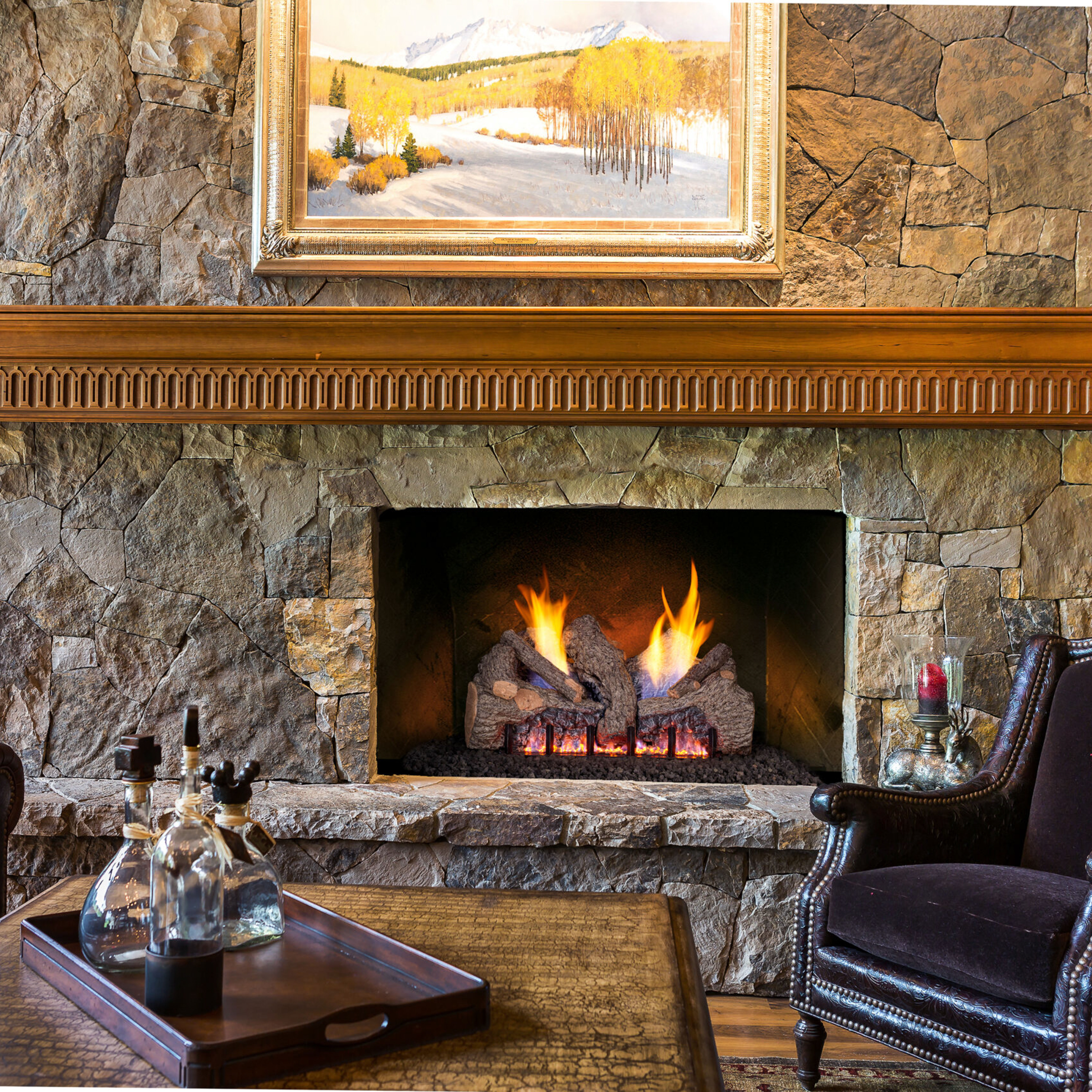 A rustic, traditional living room with a velvet decorative chair and wooden accents, along with a large, stone fireplace, a wooden mantel, and a Ventless gas log set.