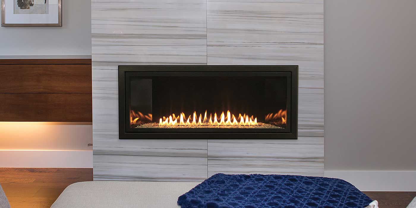 The Empire Contemporary Boulevard Ventless Gas Fireplace installed on a white, textured wall with a grey, geometric rug, a white ottoman and a blue accent blanket.