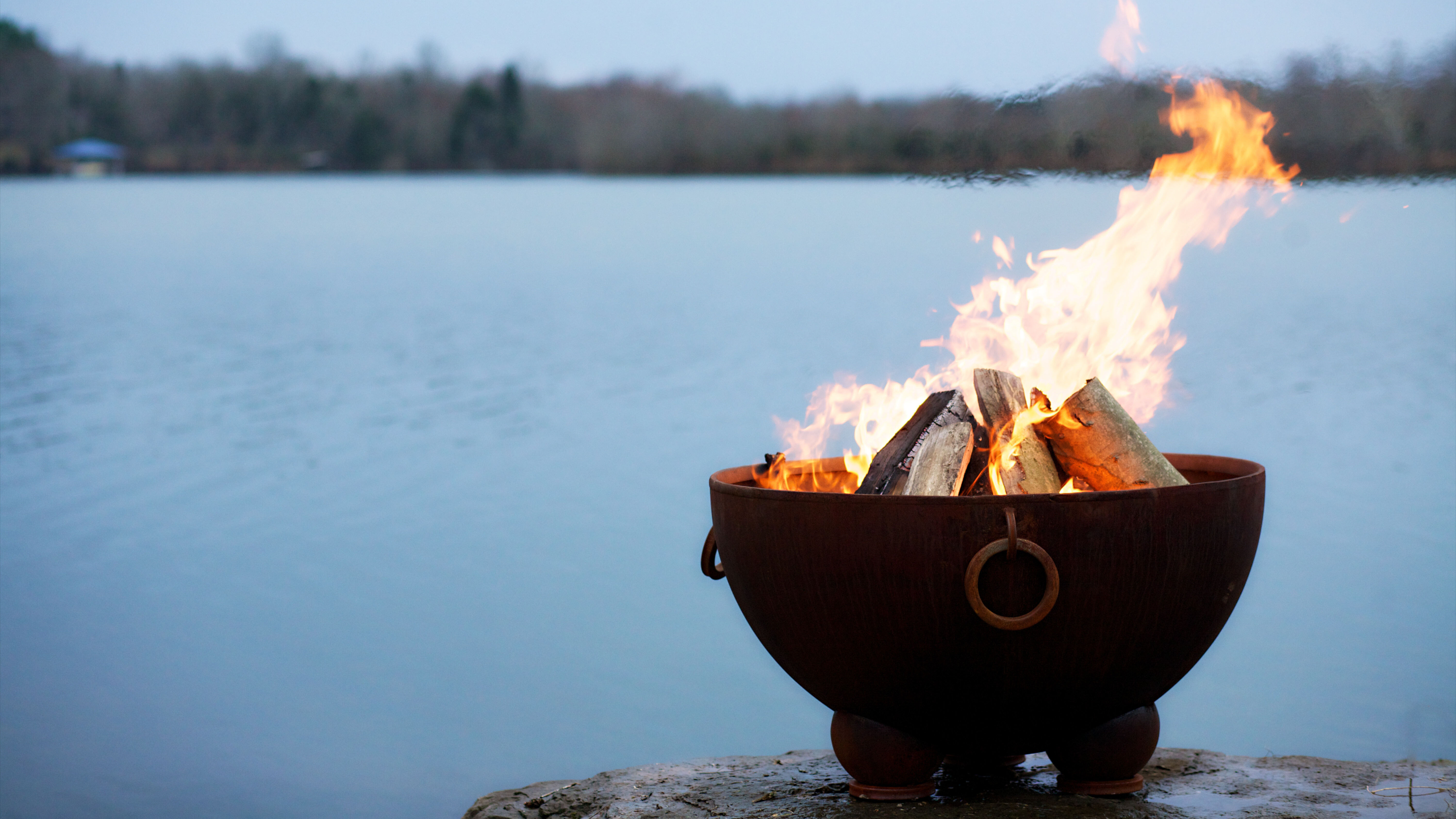 A small, steel wood burning fire pit sitting on the edge of a rock with a lake view in the background.