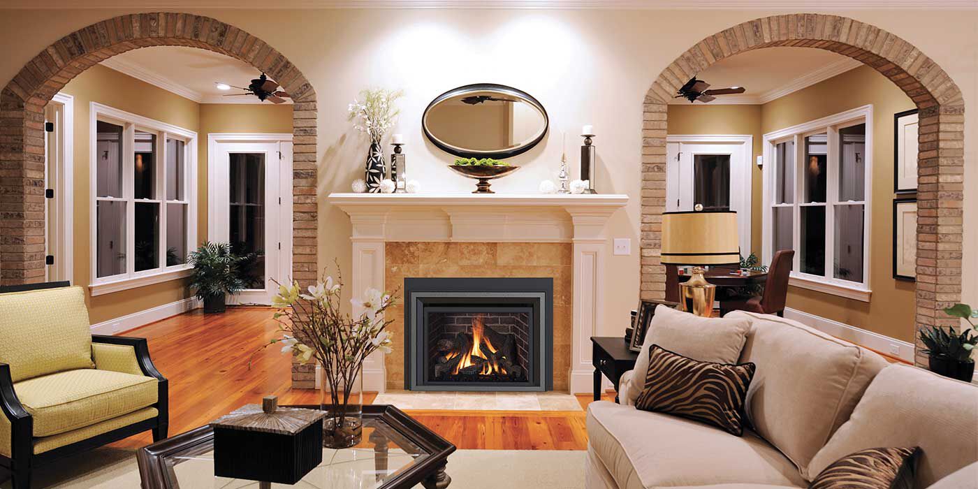 A traditional living room with two arched entryways to the living room, a large beige couch and accent chair, wooden and glass coffee and side tables, and a hearth with a white mantel and a square direct vent gas fireplace insert.