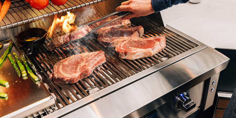 A close-up shot of several thick bone-in ribeye steaks grilling on a stainless steel grate. A flare-up of orange flames licks the side of one steak as a person uses long metal tongs to flip it. To the left, asparagus spears and a small cast-iron melting pot sit on a flat griddle attachment, with carrots and tomatoes visible on a warming rack above.