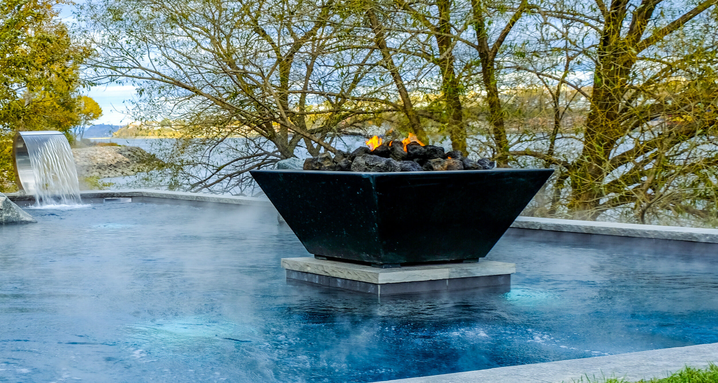 An outdoor pool with a small waterfall feature and a medium-sized concrete fire-and-water bowl burning on a platform.