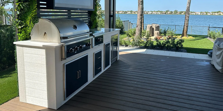A built-in stainless steel grill and outdoor kitchen station situated on a dark wood deck. The station features white cabinetry with dark grey drawers and an integrated outdoor refrigerator. A vertical green wall and a television mounted on a slat fence are behind the grill. In the background, a lawn with palm trees overlooks a large body of water lined with houses.