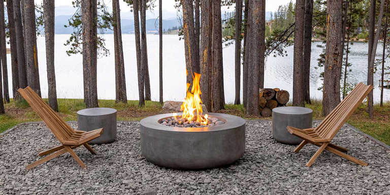 A rustic outdoor campfire in a wooded area at the lake with two wooden lounge chairs, two concrete stools, and a large concrete gas fire pit.