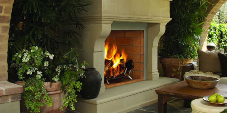Superior's Outdoor Wood Burning Fireplace installed with a traditional-looking, white hearth, a brick firebox, and a large smoke dome, surrounded by potted plants and flowers.