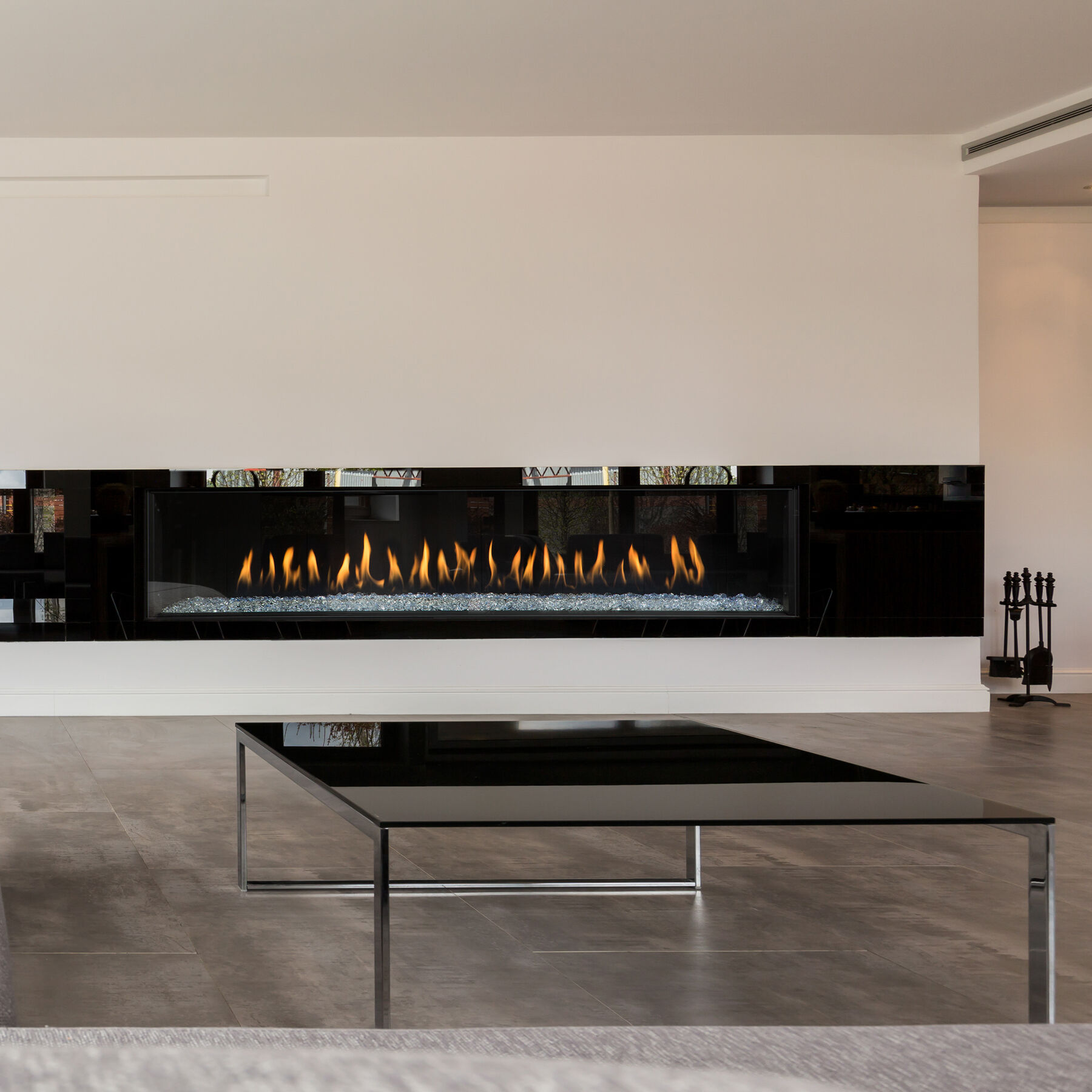 A modern, minimalist living room with a large black metal coffee table, dark hardwood flooring, white walls, and an extra-large, linear Prodigy Series gas fireplace from Montigo.