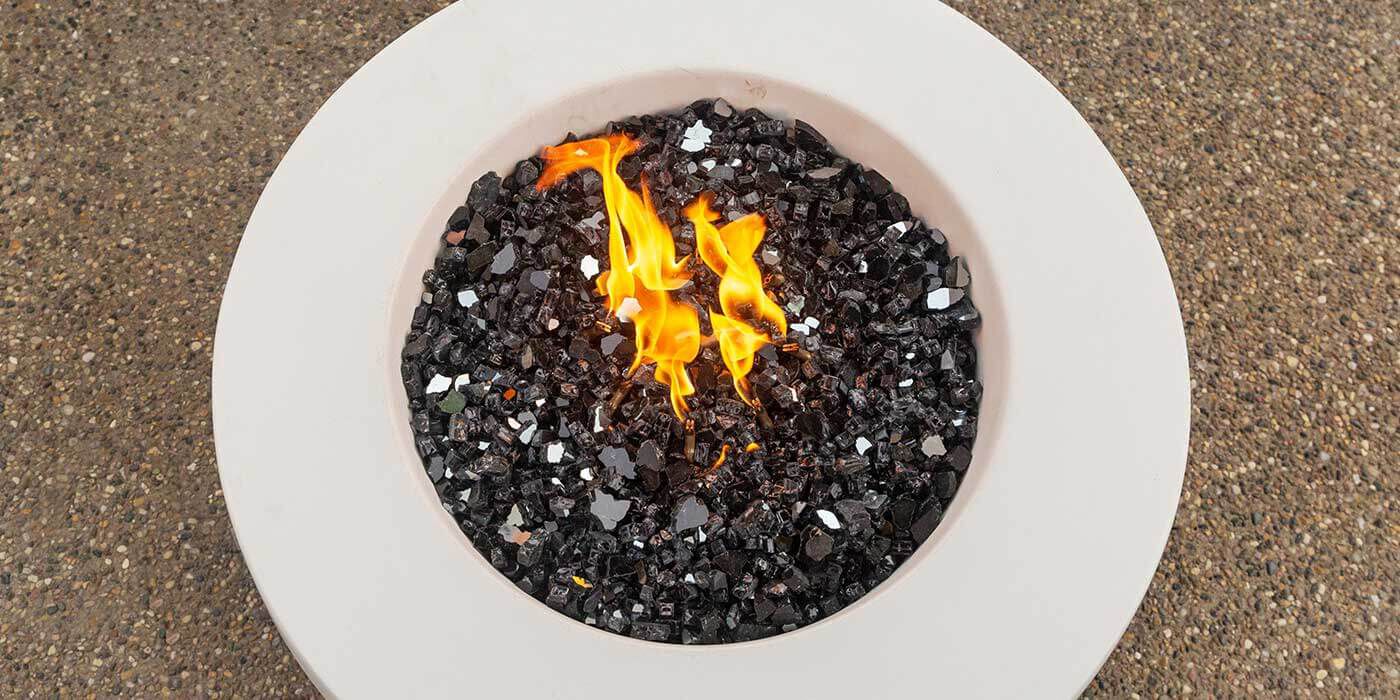 An overhead view of a white, round gas fire pit with black reflective fire glass media.