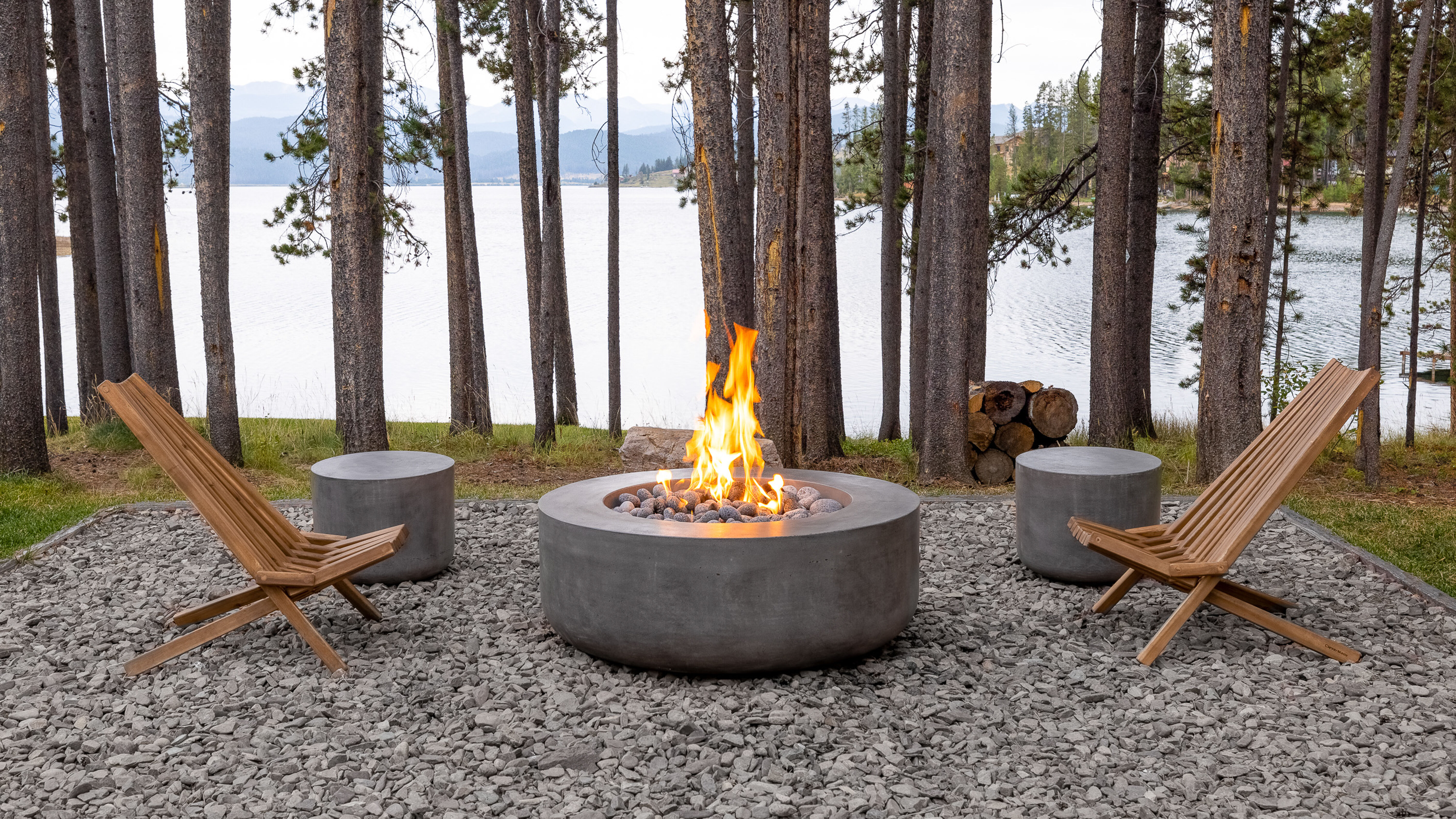 A rustic outdoor campfire in a wooded area at the lake with two wooden lounge chairs, two concrete stools, and a large concrete gas fire pit.