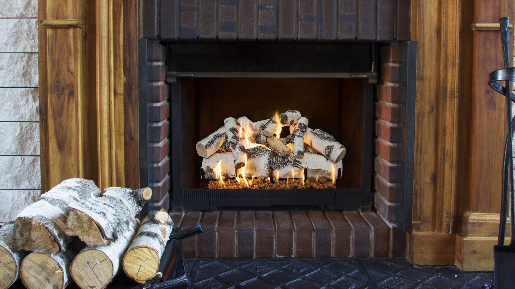 A set of Birch wood gas logs installed in a traditional masonry hearth with a wooden mantel and black fireplace tools.