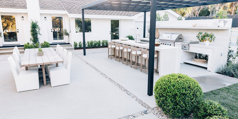 A wide-angle shot of a modern, white outdoor kitchen and dining area on a concrete patio. The space features a built-in stainless steel grill and side burner, a long waterfall island with a sink and six barstools under a black pergola, and a separate wooden dining table with eight white chairs. The background shows a white ranch-style house with black-trimmed doors and manicured landscaping.