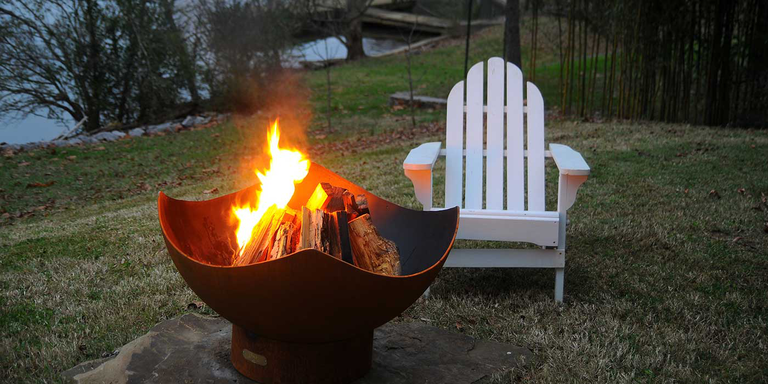 An outdoor wood burning fire pit in a backyard next to a white lounge chair