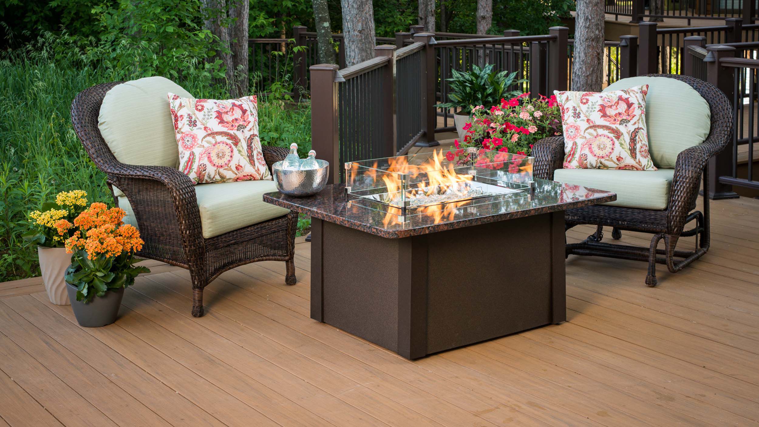 A gas fire table installed on a wooden deck in a residential backyard with white and dark wood lounge chairs, plants, and potted flowers.