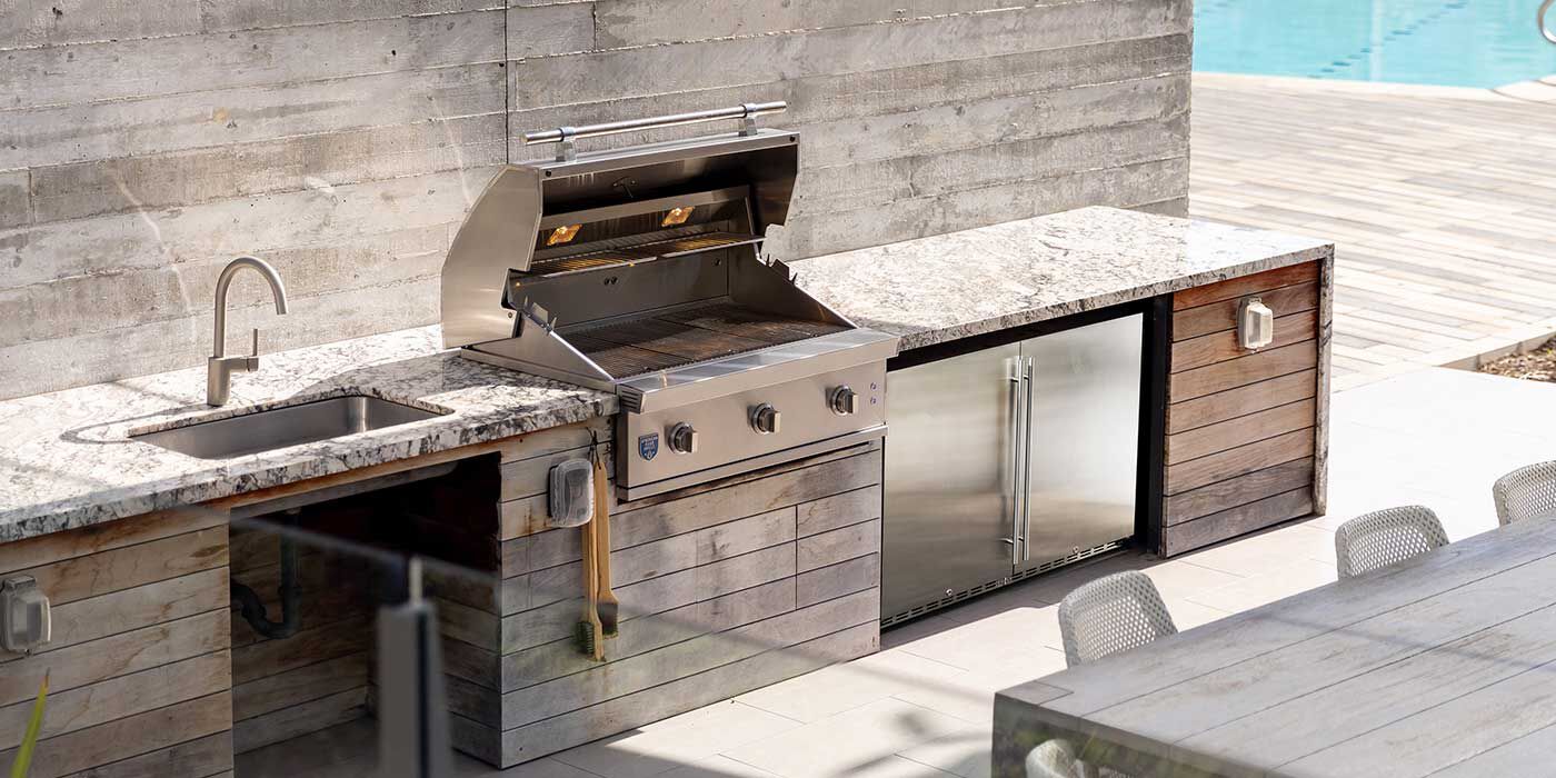 An outdoor kitchen island with a built-in, stainless steel grill, a refrigerator, and a sink with a faucet.