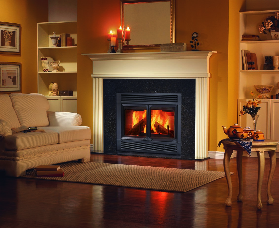 A traditional living room with shiny, dark hardwood flooring, a plush, beige couch, built-in shelves with photos, an ornate, white side table, and a large hearth with a white mantel and a black wood burning fireplace.