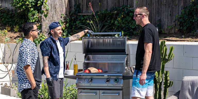 A group of men gathered around an open gas grill outside on the patio.