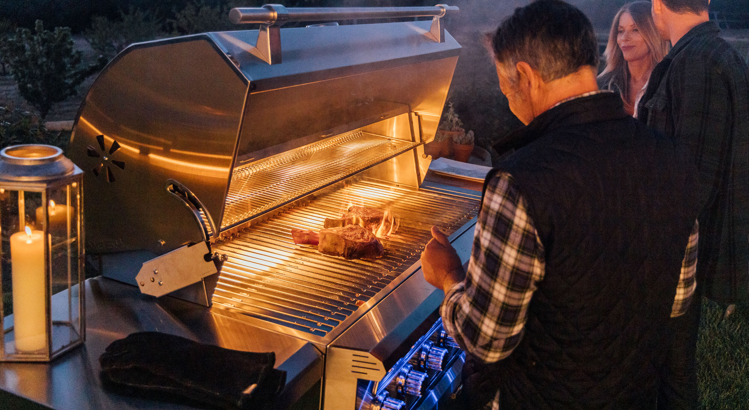 Three people gathered around a stainless steel grill at night, admiring two large steaks sizzling on the grates.