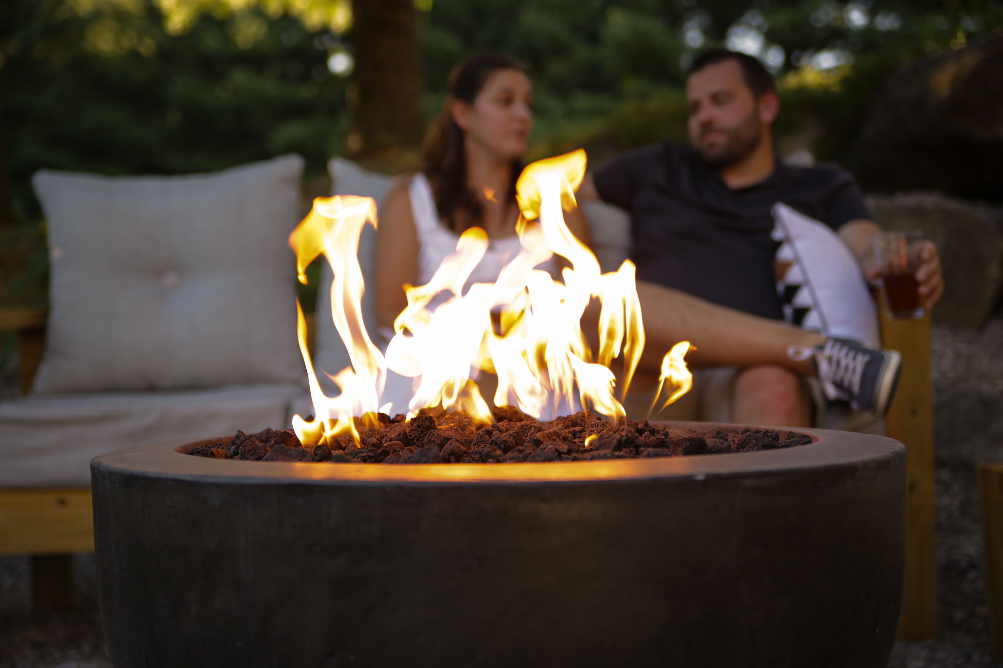 An outdoor lounge area with a small gas fire bowl in focus and a couple laughing together on a couch in the background.