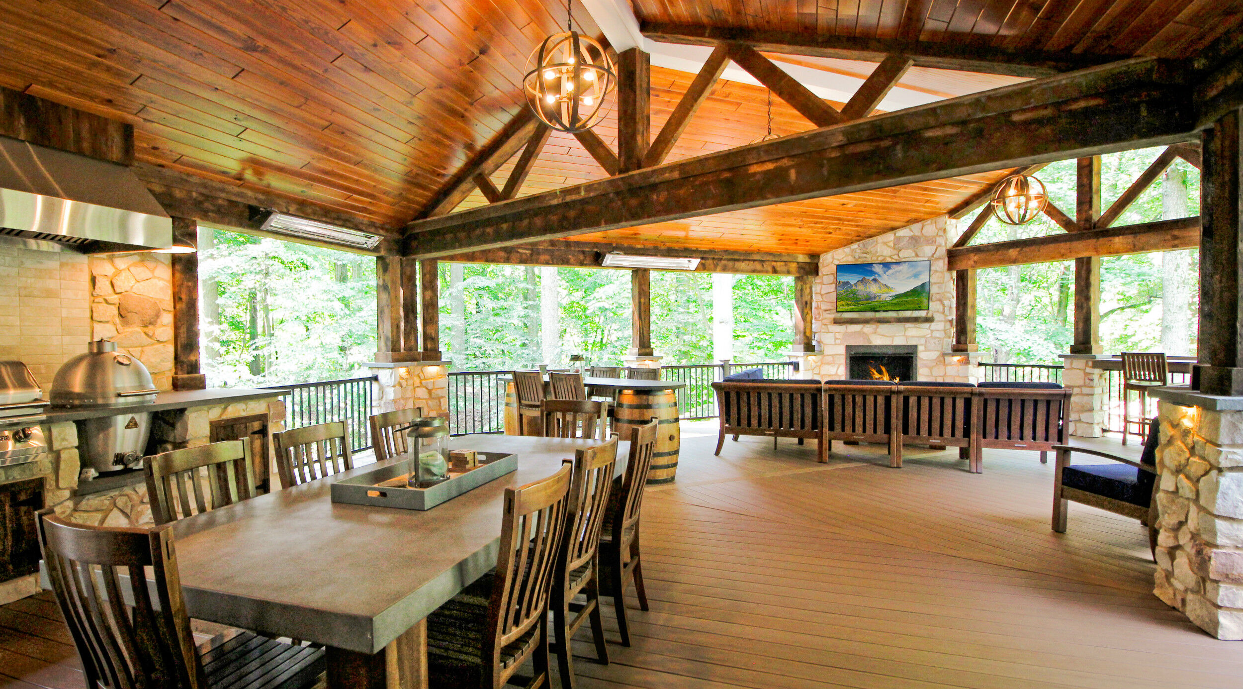 A rustic outdoor patio space with wooden walls and ceilings, a large wooden dining table and matching chairs, a lounge area with a blue couch and chairs, and a natural stone hearth with a flatscreen TV and a traditional-looking Kingsman outdoor gas fireplace.