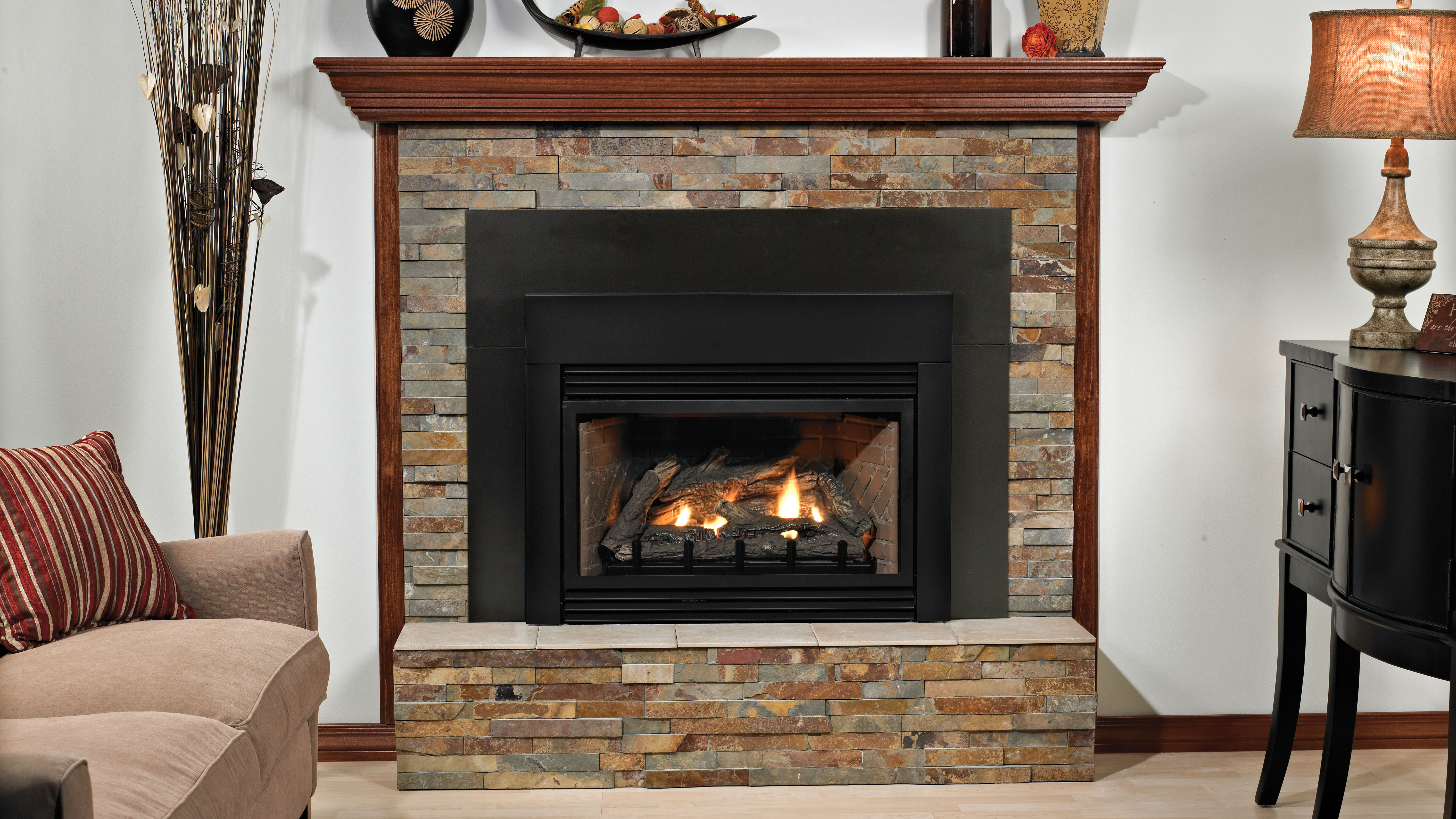 A traditional living room with a beige couch, a black buffet table with a decorative lamp, and a large, natural stone hearth with a dark wood mantel, and a rustic, wood burning fireplace.