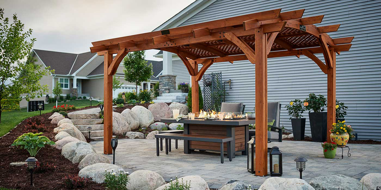 A gas fire pit table, dining bench, and dining chairs underneath a wooden pergola in a backyard