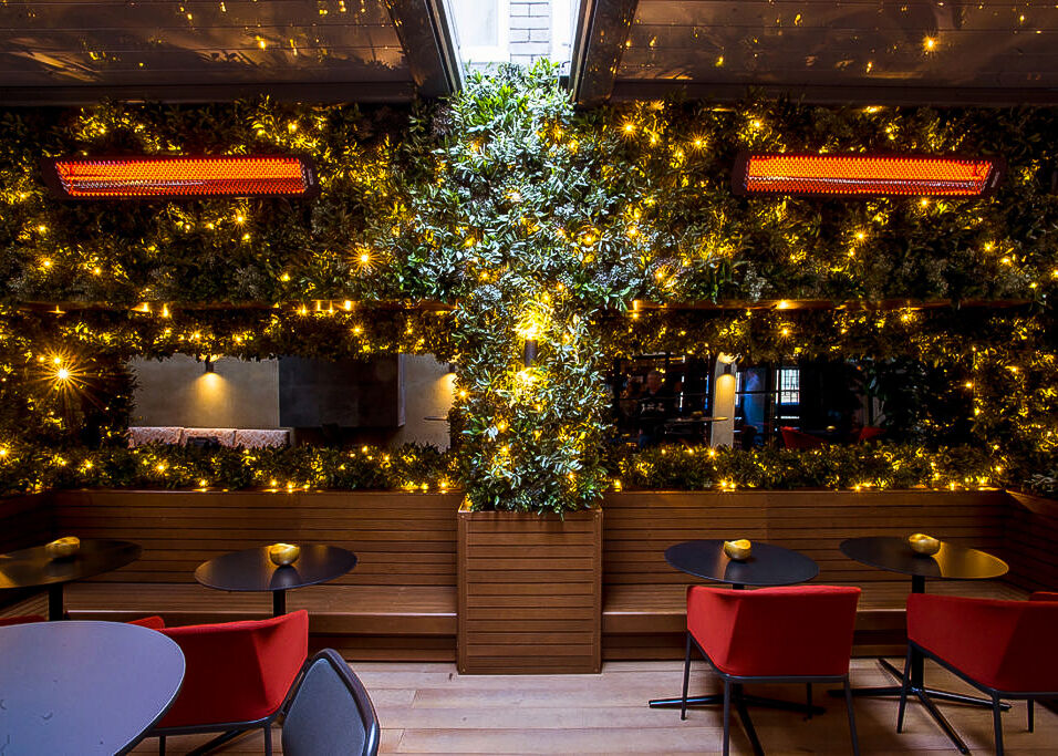 An outdoor restaurant patio with two tables, red chairs, greenery and fairy lights on the ceiling, and two overhead gas patio heaters.