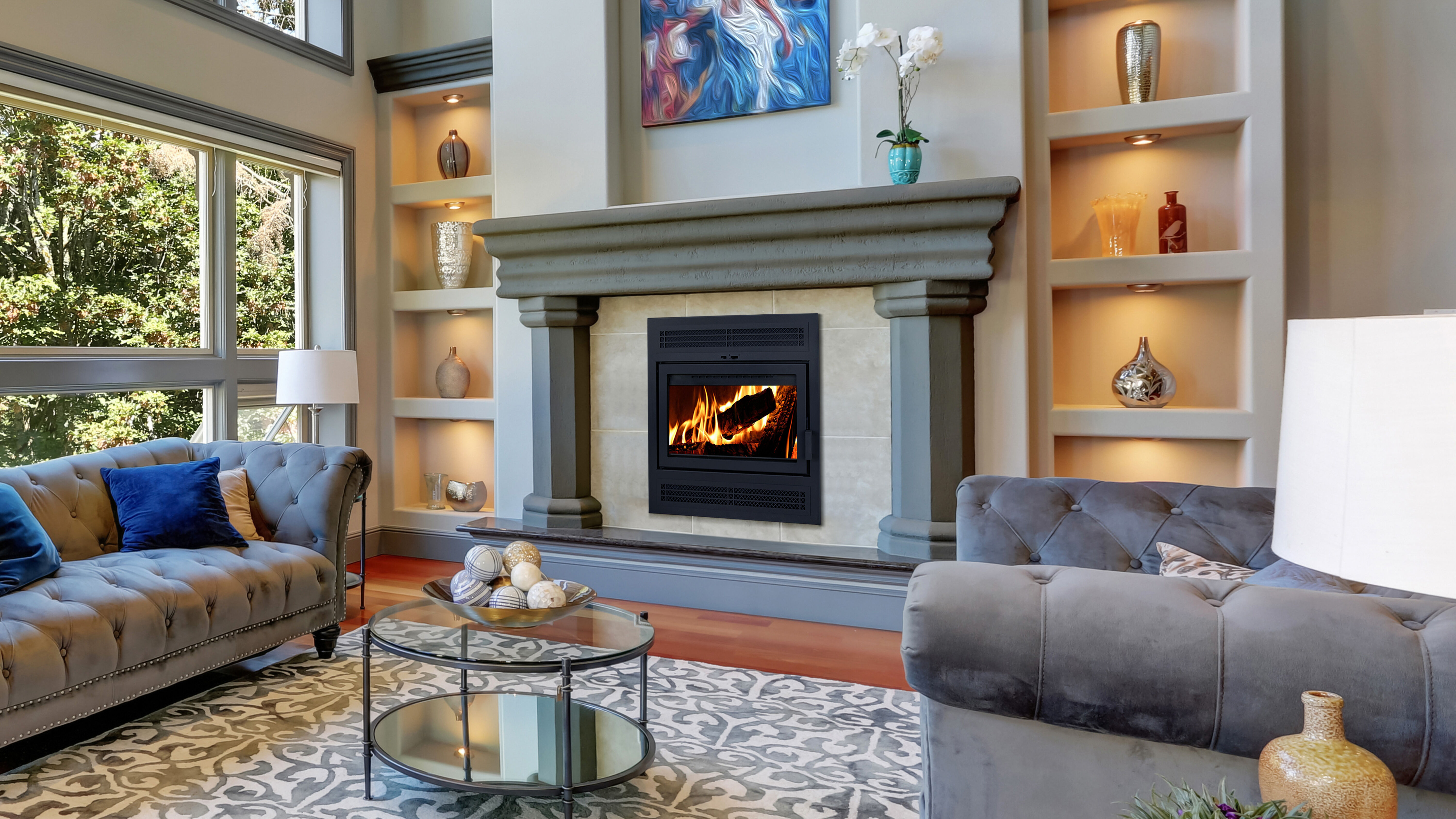 A traditional living room with gray couches, blue accent pillows, a small round glass coffee table, and a large hearth with a dusty blue mantel and a Supreme Astra traditional wood fireplace.