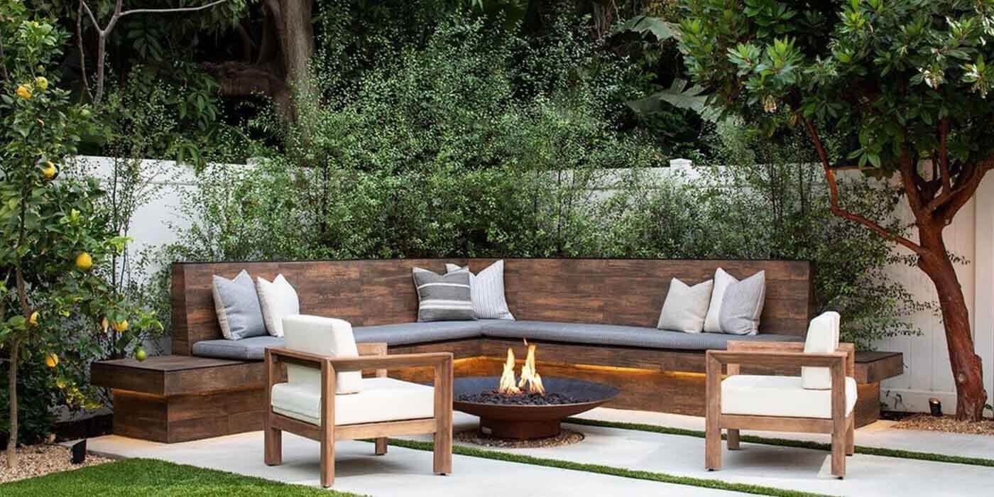 A custom-built seating and fire pit area in a contemporary backyard with a wall of lush greenery and a metal gas fire bowl with an aged patina finish
