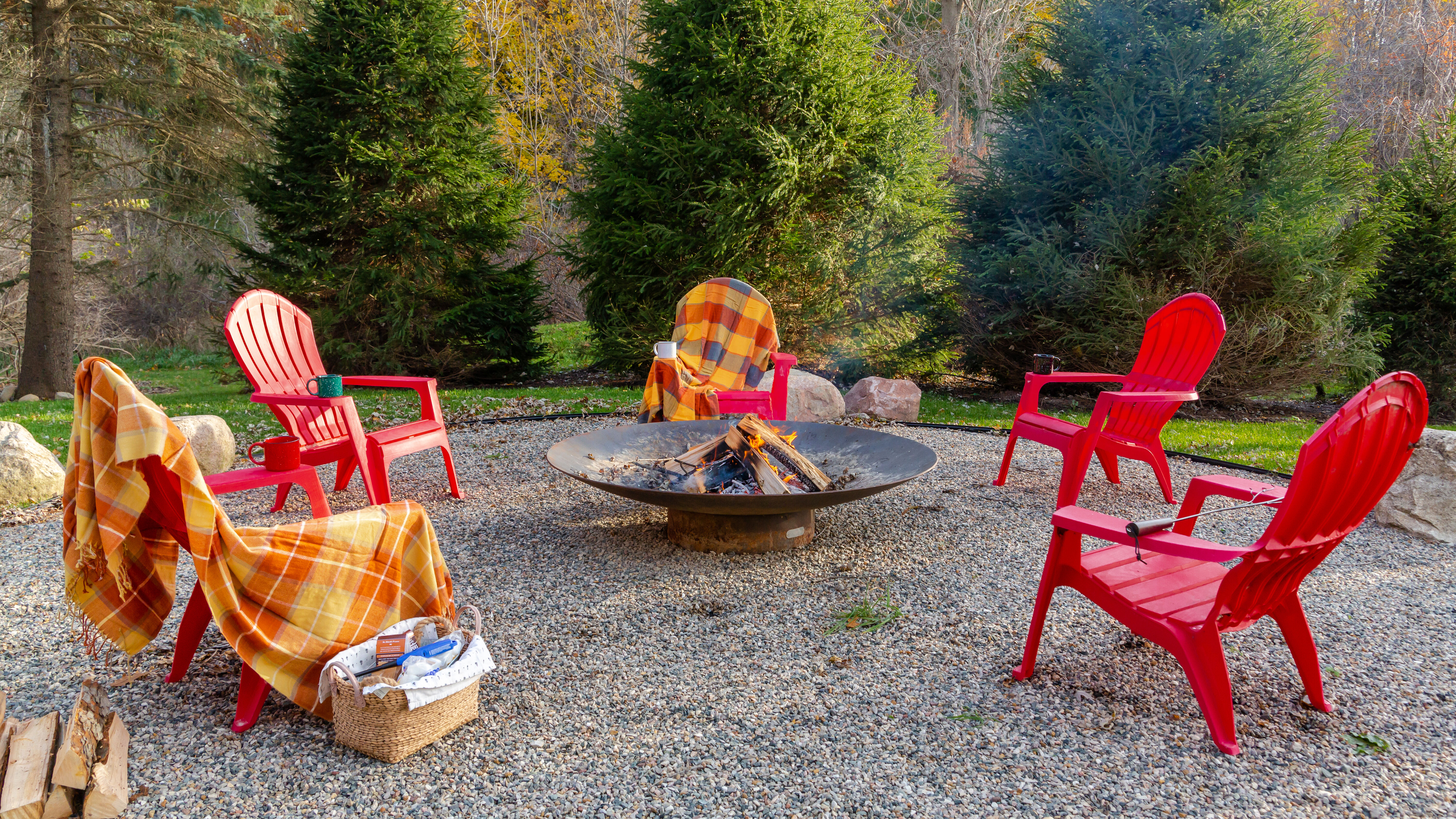 A rustic outdoor bonfire space in the woods with five bright red plastic lounge chairs, orange and yellow plaid blankets, mugs of hot cocoa, and a large steel wood burning fire pit in the center.