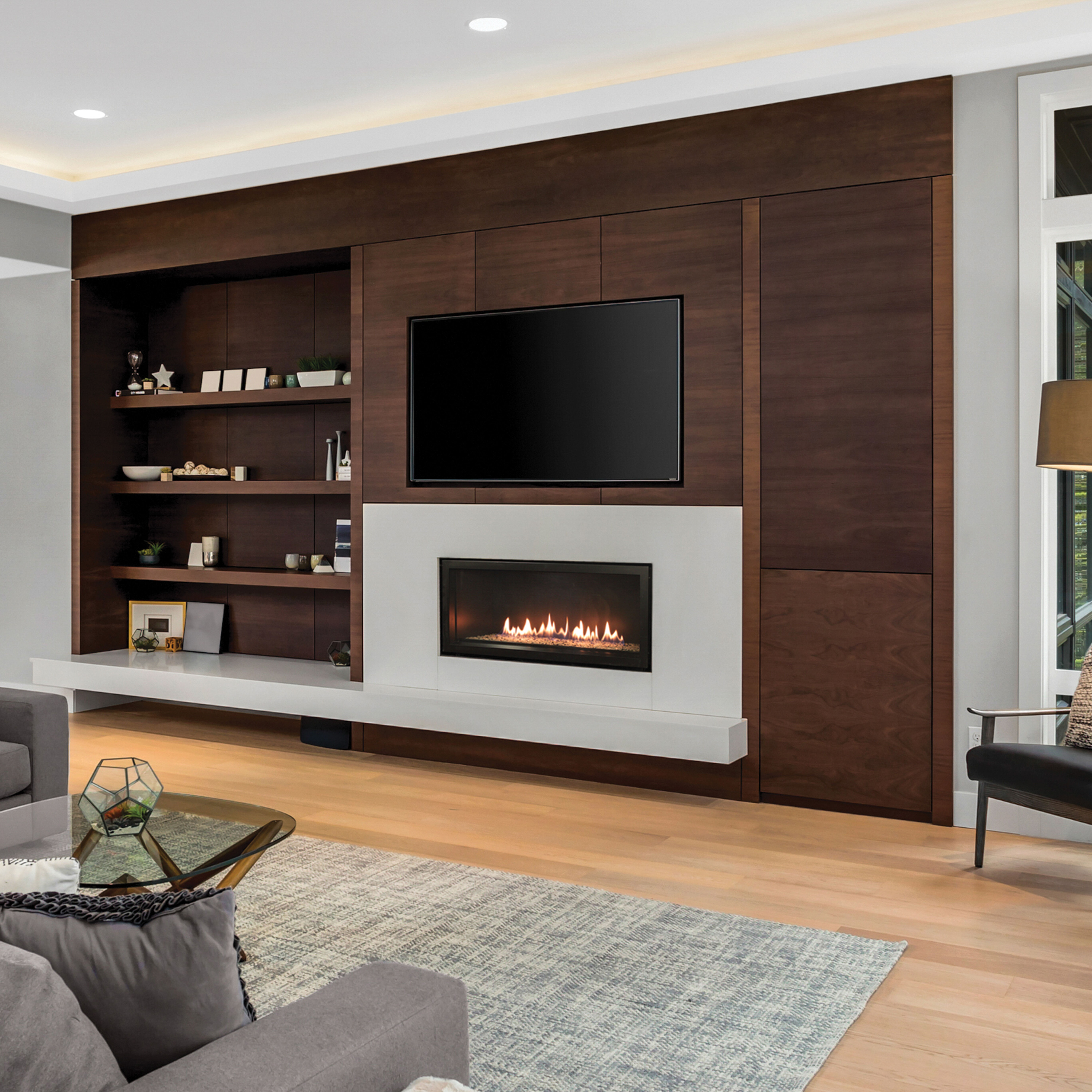 The Empire Contemporary Boulevard Direct Vent Gas Fireplace installed into an entertainment center in a living room with brown shelves and a large TV mounted over the fireplace.