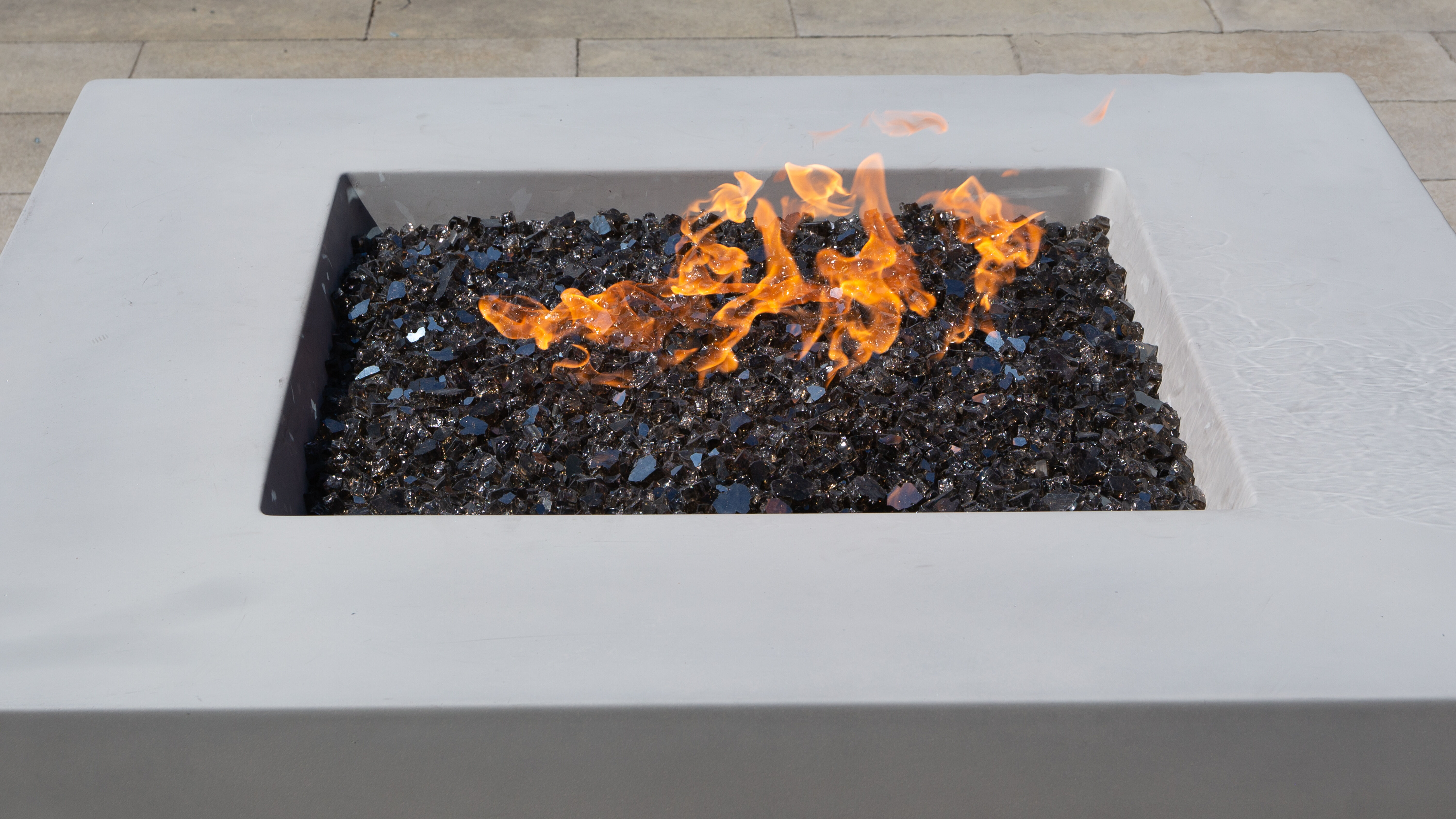 An aerial view of a light gray Quadro gas fire pit by FlameCraft with black reflective fire glass media and orange flames.