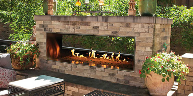 The Carol Rose Linear See-Through Fireplace by Empire install outside on an open patio, finished with neutral-colored bricks and surrounded by green shrubs, trees, plants, and potted flowers.