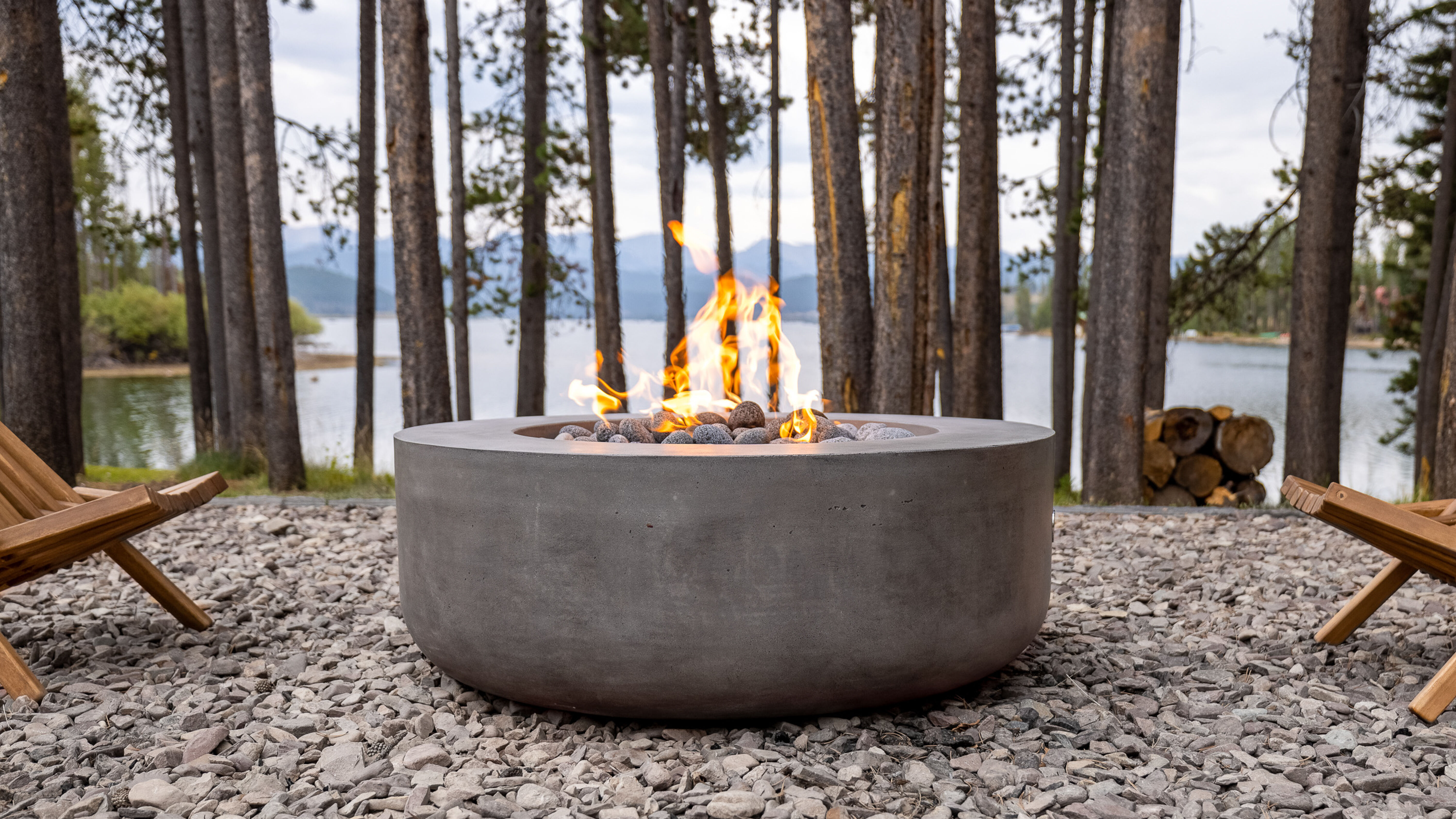 An outdoor lounge area with tall trees and a lake in the background, two wooden lounge chairs, and a large, concrete gas fire pit with rolled lava stones.
