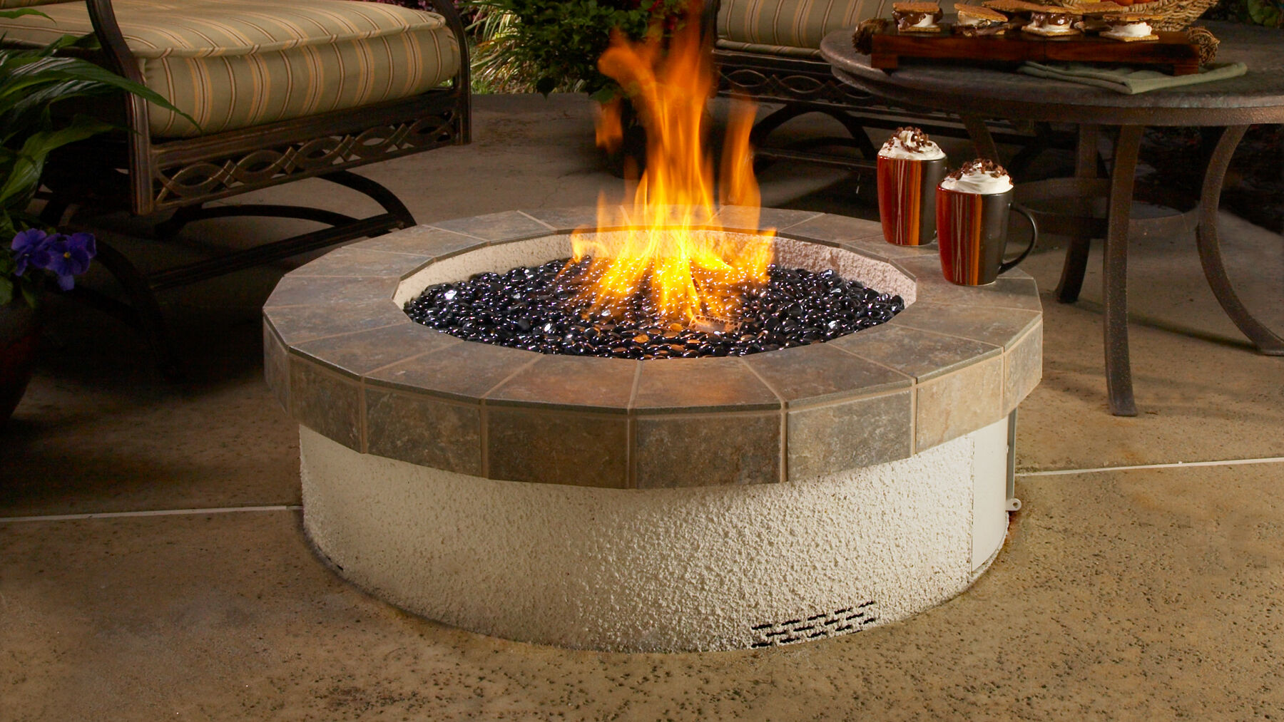 An outdoor patio space with a lounge chair and a large stucco and stone gas fire pit with smooth black fire stones.